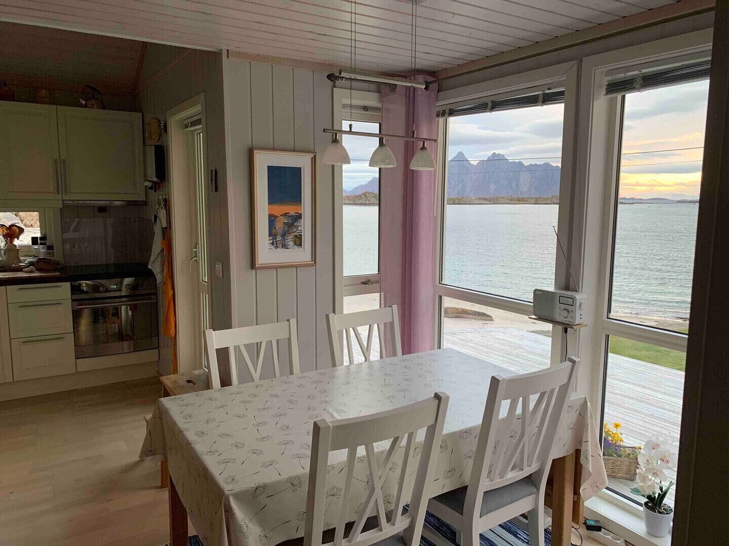 Ein Essbereich mit Tisch und Stühlen, eine Küche im Hintergrund und Blick auf ein Gewässer mit Bergen.