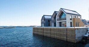 Ein modernes Haus mit einer Holzterrasse, die über dem Wasser liegt, großen Fenstern und einem geneigten Dach.