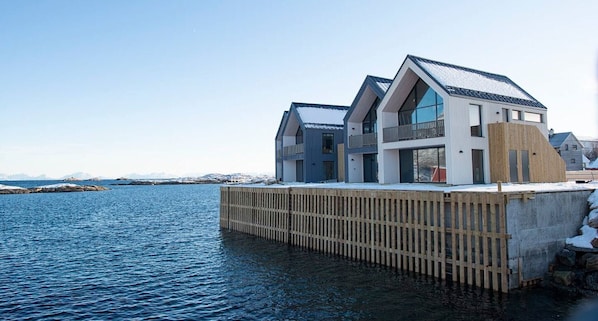 A modern house with a wooden deck extending over water, large windows, and a sloped roof.
