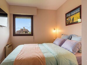 A bedroom with a bed, a window with a view of buildings, a wall-mounted light, and a framed picture. - Les Manoirs by Interhome (Deauville-Trouville)