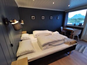 A modern bedroom with a bed, bedside lamps, a desk, and a view of a landscape. - Panoramic seaview cabin for two persons by the sea (Flakstad)