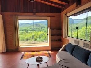 Une pièce dotée d’une grande fenêtre donnant sur des collines verdoyantes, d’un canapé, d’une petite table et d’un tapis à motifs.