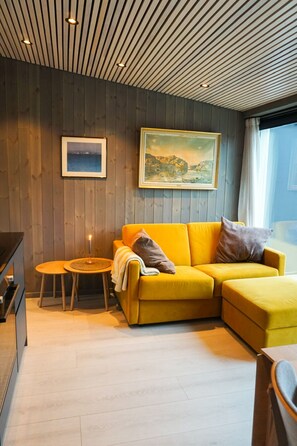 Ein modernes Wohnzimmer mit Holzboden, einem gelben Sofa, einem kleinen runden Tisch und zwei gerahmten Bildern an der Wand.