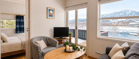 A cozy living room with a sofa, a grey armchair, a wooden coffee table, a TV, and a large window with a view of a snowy landscape.