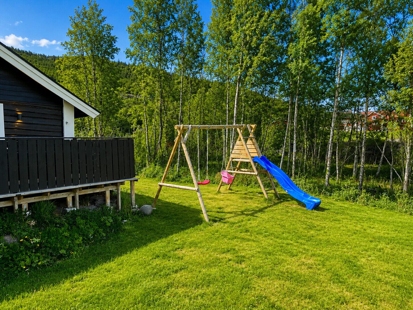 A wooden swing set with a blue slide in a grassy backyard with a wooden fence and a building in the background.