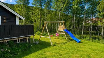 A wooden swing set with a blue slide in a grassy backyard with a wooden fence and a building in the background.