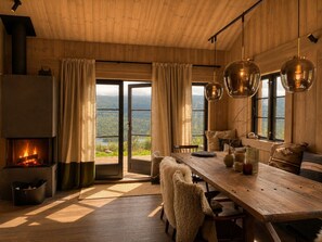 Une pièce chaleureuse avec une cheminée, une grande table en bois et une vue sur un paysage boisé par les fenêtres.