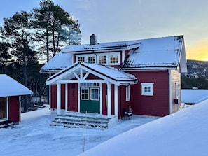 Une maison rouge à deux étages, avec une porte verte et des boiseries blanches, entourée d’un paysage enneigé et d’arbres.