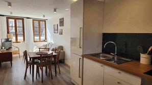 A modern kitchen with a wooden countertop, stainless steel sink, and white cabinetry. There is a dining area with a table and chairs, and a television on a desk.