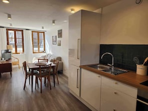 A modern kitchen with a wooden countertop, stainless steel sink, and white cabinetry. There is a dining area with a table and chairs, and a television on a desk.