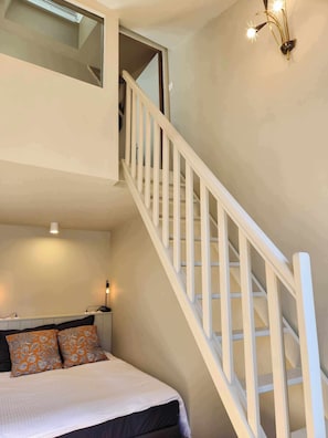 A bedroom with a bed, a staircase, a nightstand, and a wall-mounted light fixture.
