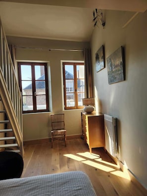 A room with a bed, a chair, a dresser, two windows, a radiator, and a staircase.