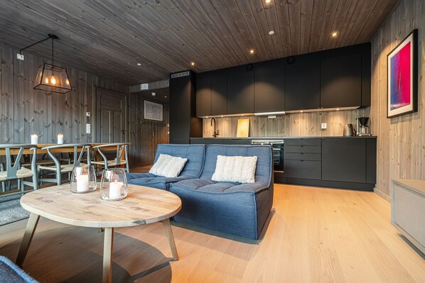 Ein modernes Interieur mit Holzboden, einem blauen Sofa mit weißen Kissen, einem runden Holztisch mit Kerzen und einer Küchenzeile mit dunklen Schränken.