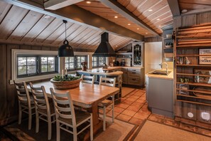 A kitchen with a wooden ceiling, a dining table with chairs, a black pendant light, and a view through the windows.