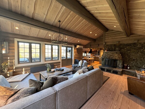A log cabin interior with a stone fireplace, wooden ceiling beams, and a large window overlooking a scenic landscape.