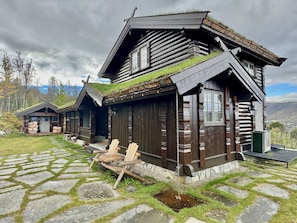 Eine Blockhütte mit einem pflasterbelagten Eingang, Holzstühlen und einem grasbewachsenen Dach.
