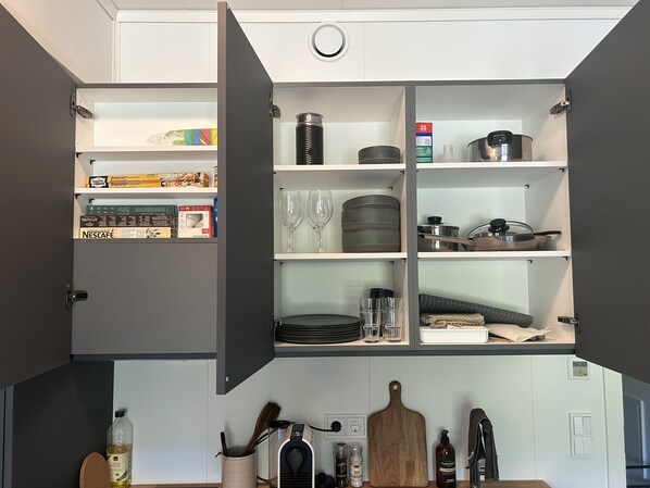 A kitchen with open cabinets displaying various kitchenware, a coffee machine, and a wooden cutting board.