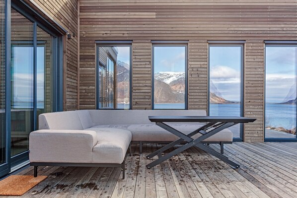Ein moderner Außenbereich mit Sitzgelegenheit, einer Couch und einem Tisch, großen Fenstern und einer Holzterrasse.