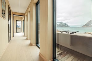 A hallway with wooden flooring and large windows, leading to a balcony with a view of a lake and mountains.
