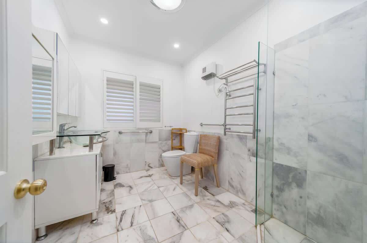A bathroom with a glass shower enclosure, a white sink, a toilet, and a chair.