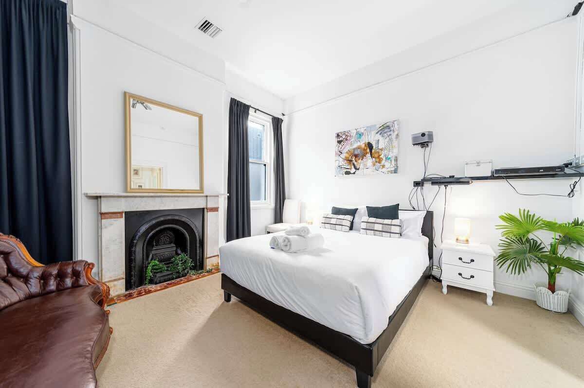 A bedroom with a bed, a fireplace, a mirror, a nightstand, a lamp, and a plant.