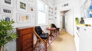 A hallway with framed pictures on the walls, a wooden dresser, two chairs, a small table with a plant, and a bookshelf with books and decorative items.