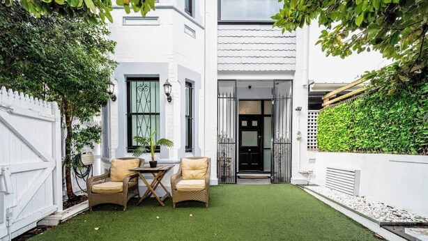 A white building with a white fence, a small lawn, two wicker chairs, and a table.