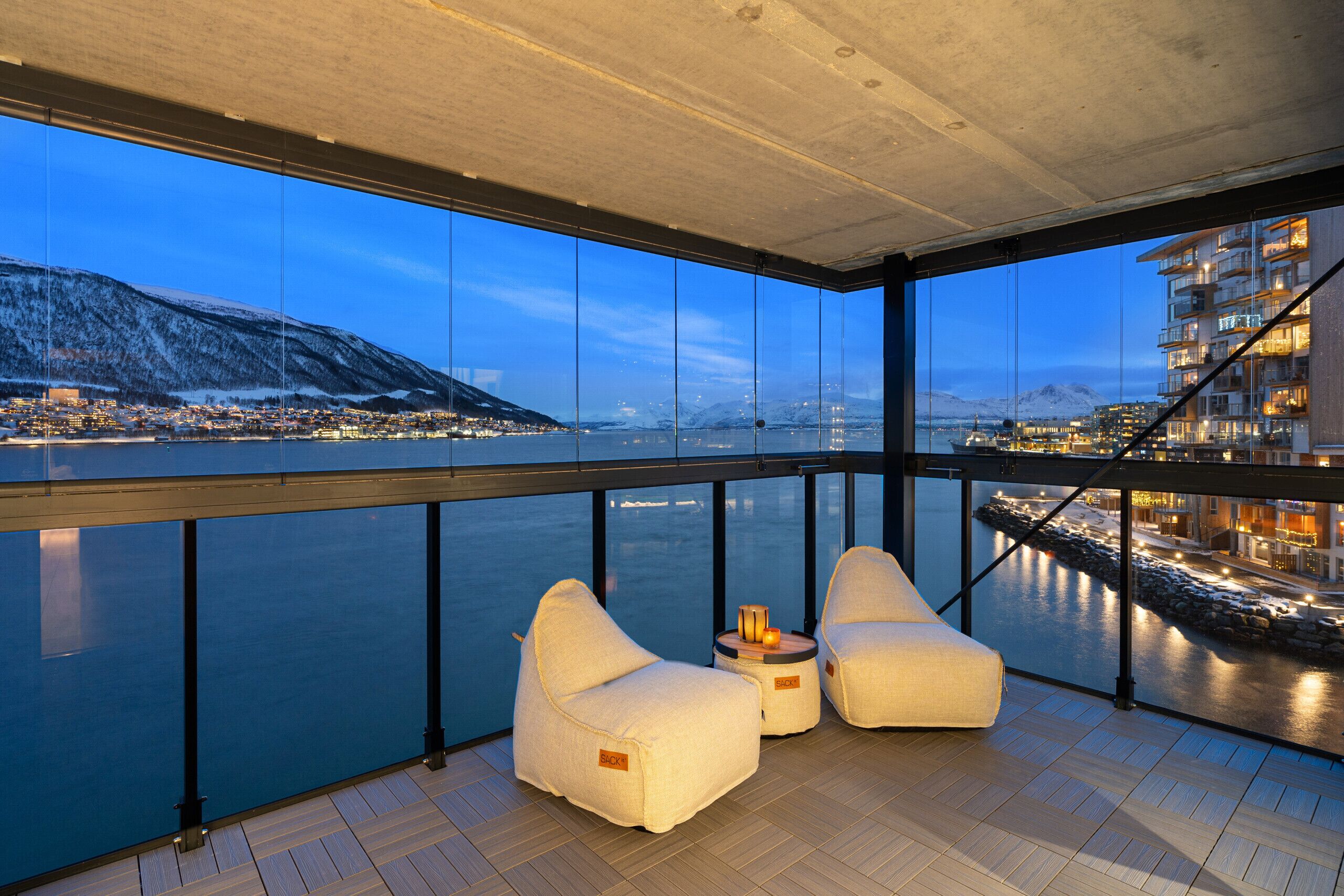 Een balkon met uitzicht op een waterpartij en een stadsgezicht, met twee witte stoelen met kussens en een tafeltje met een schaal fruit.