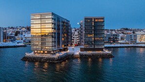 Een gebouw aan het water met een glazen gevel, gebouwd op palen boven het water, met een stadsgezicht op de achtergrond.
