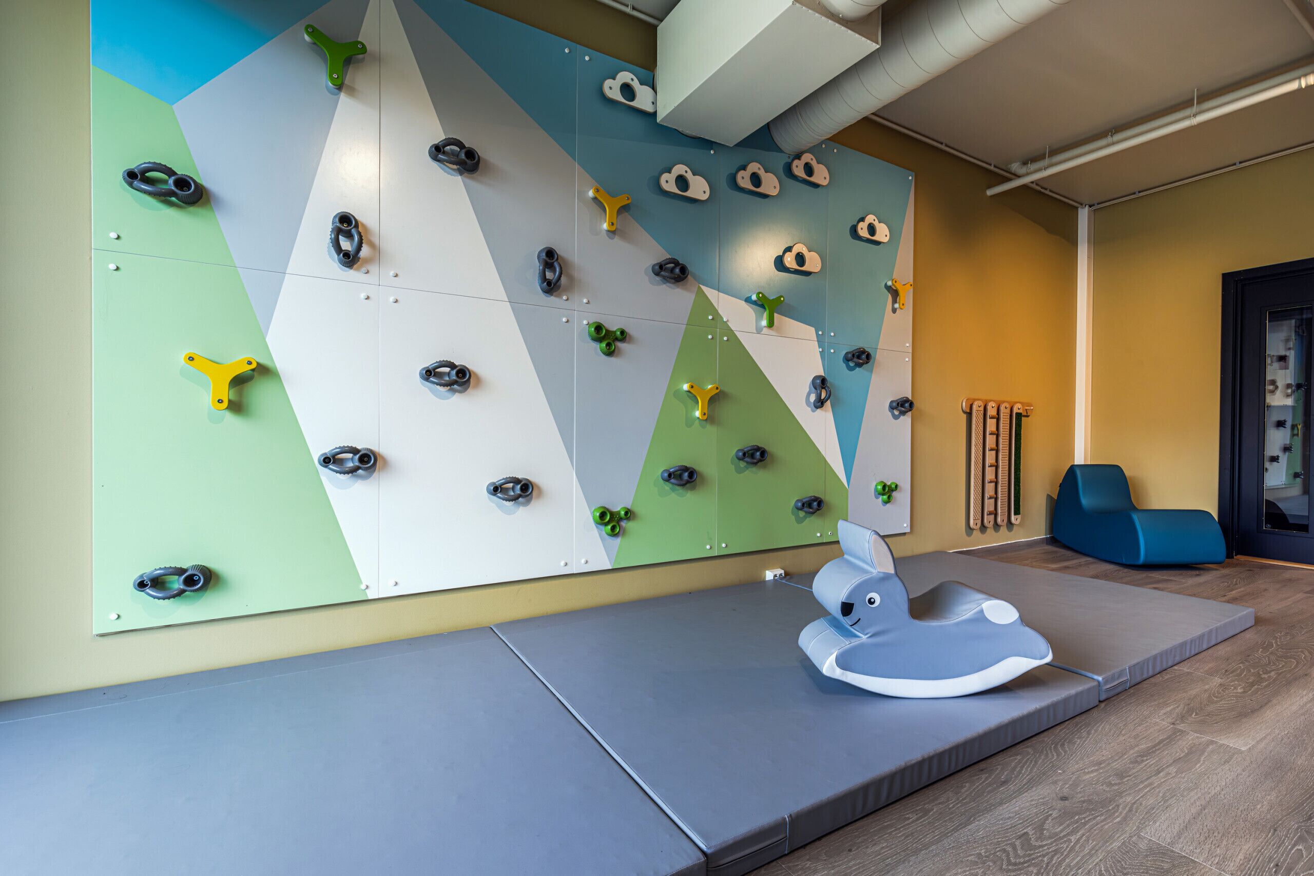 A climbing wall with various holds, a padded mat, and climbing shoes on the floor.