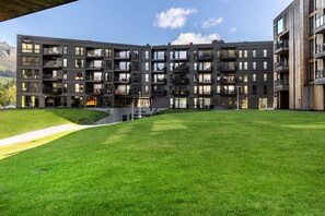 Un edifici modern de diversos pisos amb balcons, un pati amb gespa i un paisatge de muntanya al fons.