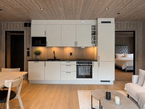 Cuina moderna amb armaris blancs, un microones negre i un forn. Hi ha una zona de menjador amb taula i cadires de fusta.