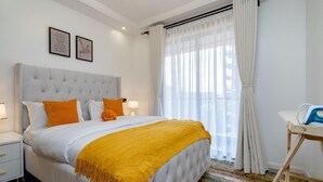 Una camera da letto ordinata con una testiera trapuntata, un letto con biancheria da letto bianco e arancione, una coperta gialla e un comodino con una lampada.