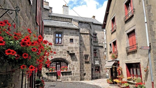 Una estrecha calle adoquinada con edificios de piedra, contraventanas rojas y plantas en macetas.