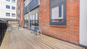 A wooden deck with a metal fence, a brick building with large windows, and a metal table and chair. - Modern 1-Bed Flat in Jewellery Quarter (West Midlands)