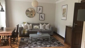 Una sala de estar con una mesa de comedor de madera, un sofá gris, alfombra estampada y un espejo redondo en la pared.