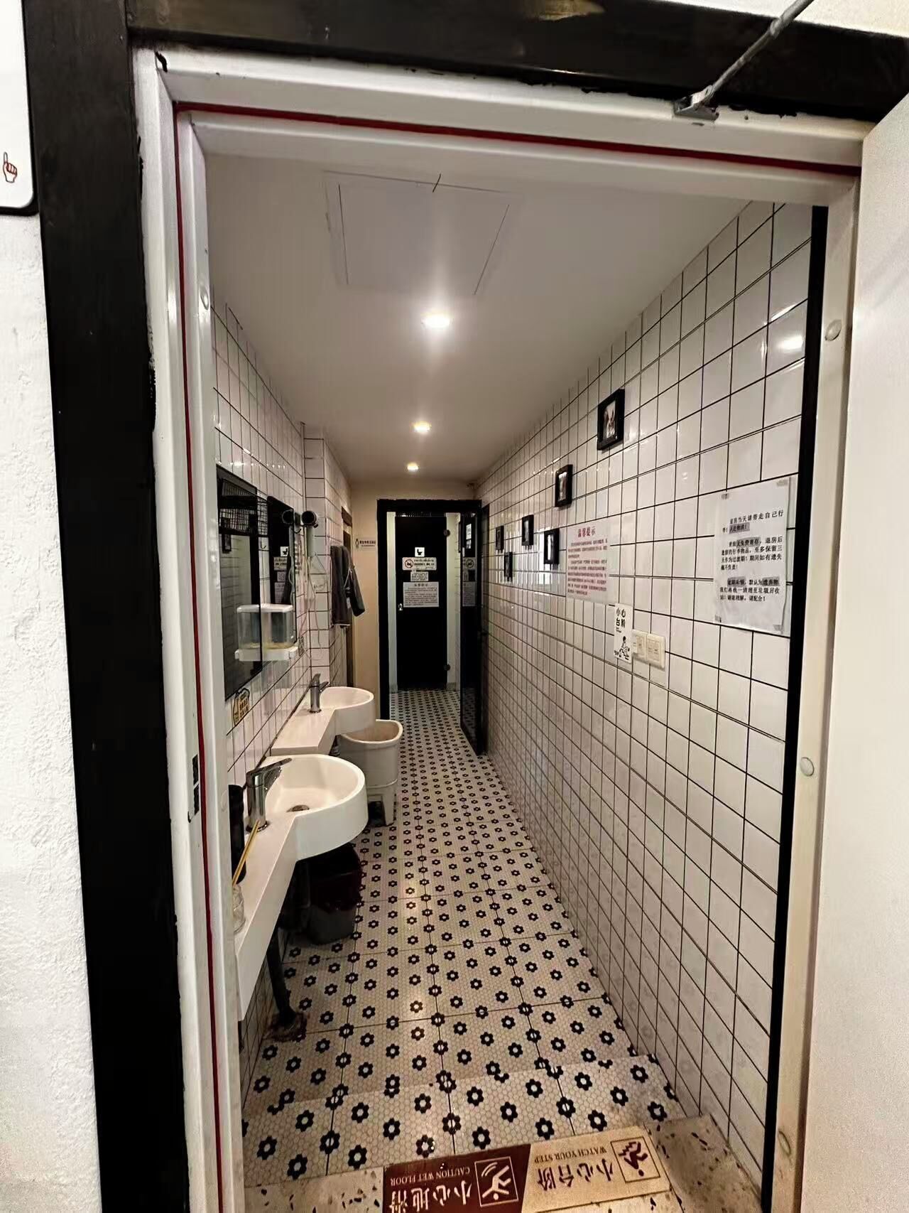 Um corredor estreito com paredes e chão em azulejo, que dá para um espaço com três pias e um espelho.