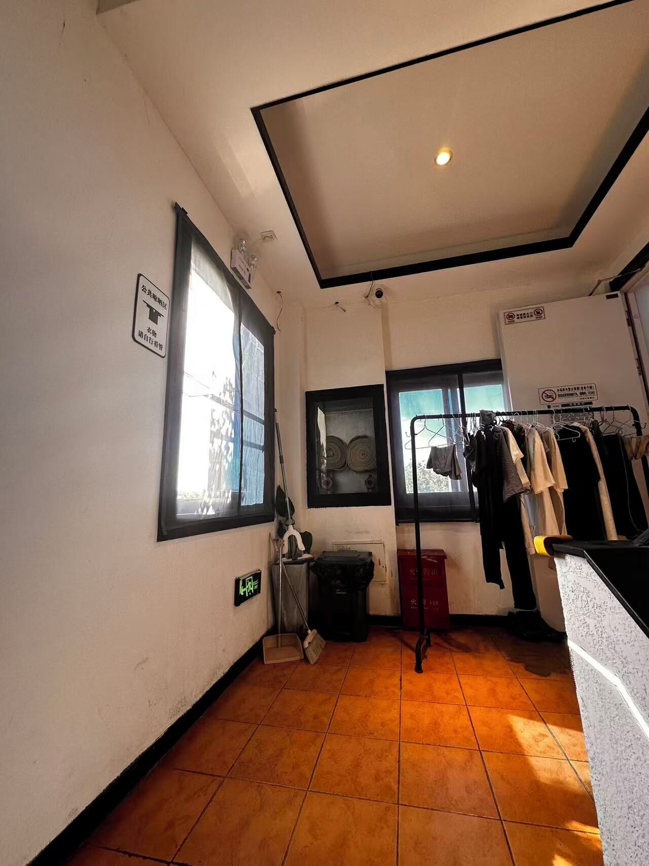 A room with a tiled floor, a window with blinds, a clothes rack with hanging garments, a trash bin, and a sign on the wall.