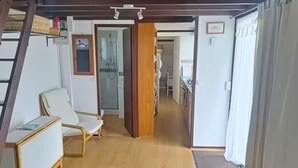 Una habitación con piso de madera, una silla blanca, una mesita y una puerta de madera. En el fondo, se observa una zona de cocina.