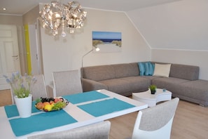 Ein modernes Wohnzimmer mit einer Couch, einem Esstisch mit Obstschale, einer Chandelierleuchtre und einem Gemälde an der Wand.