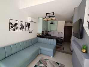Ein Wohnzimmer mit einem hellblauen Ecksofa, einer Küchenzeile inklusive Herd und Mikrowelle sowie einem flachbildfernseher an der Wand.