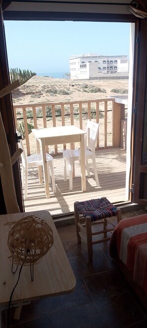 A room with a view of a sandy beach and buildings, a wooden table with chairs, a small stool, and a bed with a blanket.