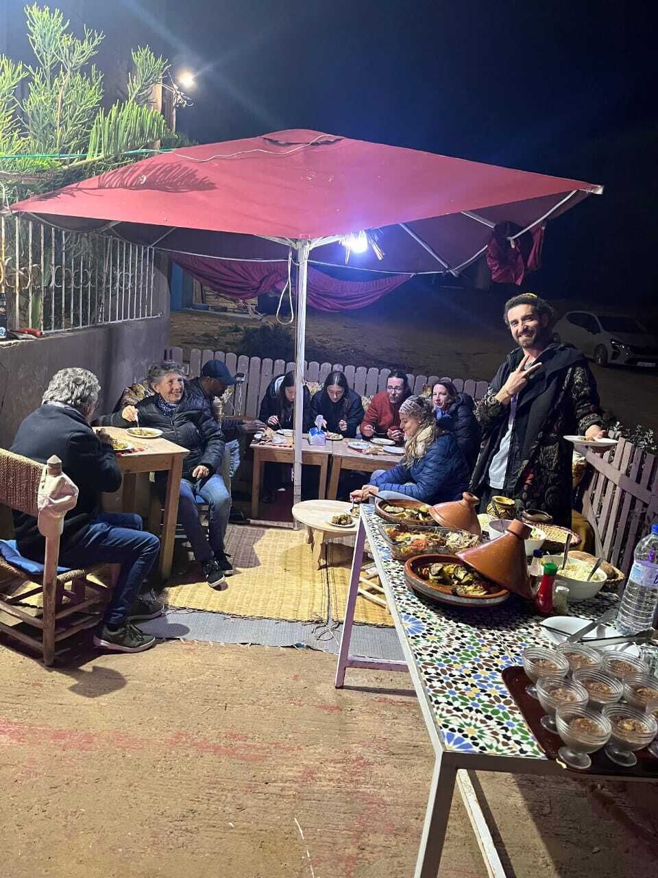Un área de comedor al aire libre con mesas, sillas y una gran sombrilla roja que da sombra.