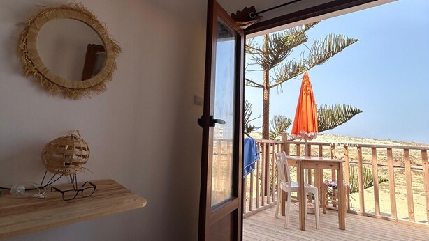 A room with a bed, a wooden shelf with a woven basket and glasses, a round mirror, and a door leading to a balcony with a wooden railing and an orange umbrella. - Tildi Surf House (Imsouane)