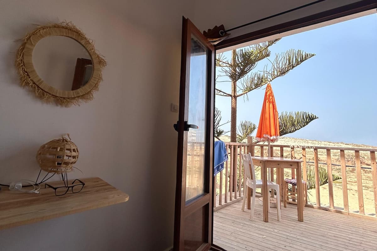 A room with a bed, a wooden shelf with a woven basket and glasses, a round mirror, and a door leading to a balcony with a wooden railing and an orange umbrella.