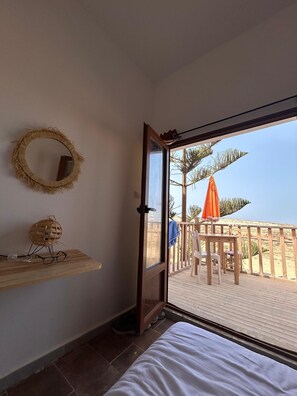 Una habitación con una cama, un estante de madera con una cesta tejida y vasos, un espejo redondo y una puerta que da a un balcón con barandilla de madera y un paraguas naranja.