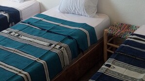 A room with two beds, each with a different patterned bedspread, white pillows, and a small stool with a colorful blanket. - Tildi Surf House (Imsouane)