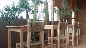 Un rincón de una habitación con una mesa y sillas de madera, el respaldo de una silla tejido, y una planta al fondo.
