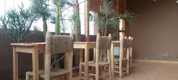 A corner of a room with a wooden table and chairs, a woven chair back, and a plant in the background.