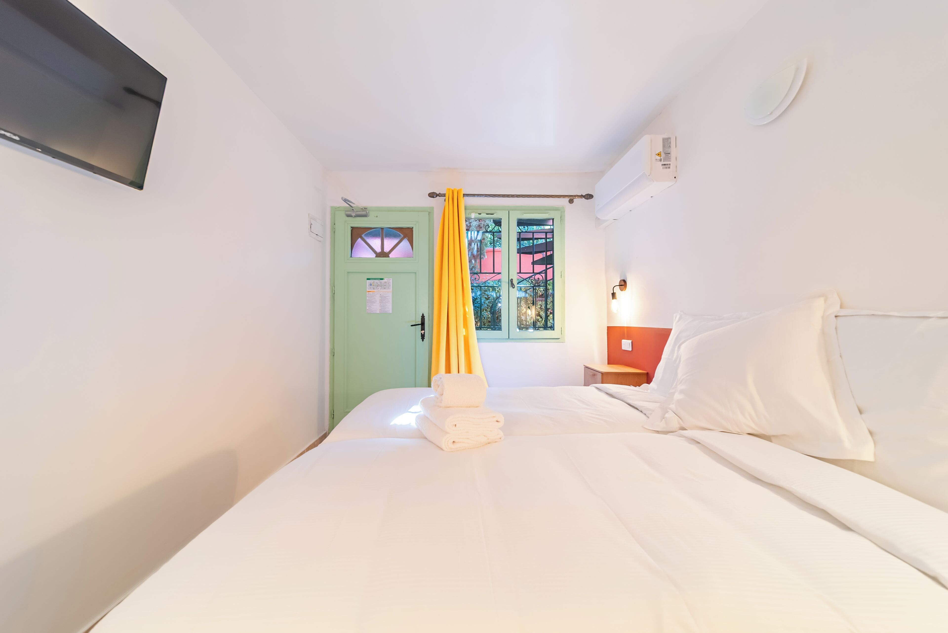 A single bed with white linens, a green door with a window, and a yellow curtain.